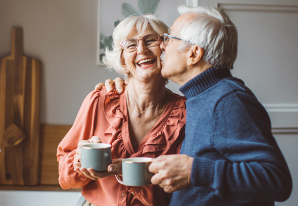 an older married couple embrace over their morning coffee as the husband kisses his laughing wife on the cheek